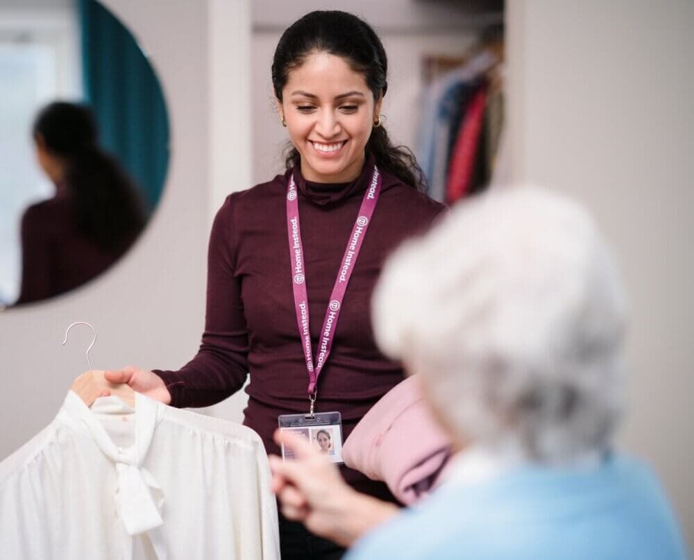 A woman with curly hair brushes the hair of an elderly person in a bright, sunlit room with large windows. - Home Instead