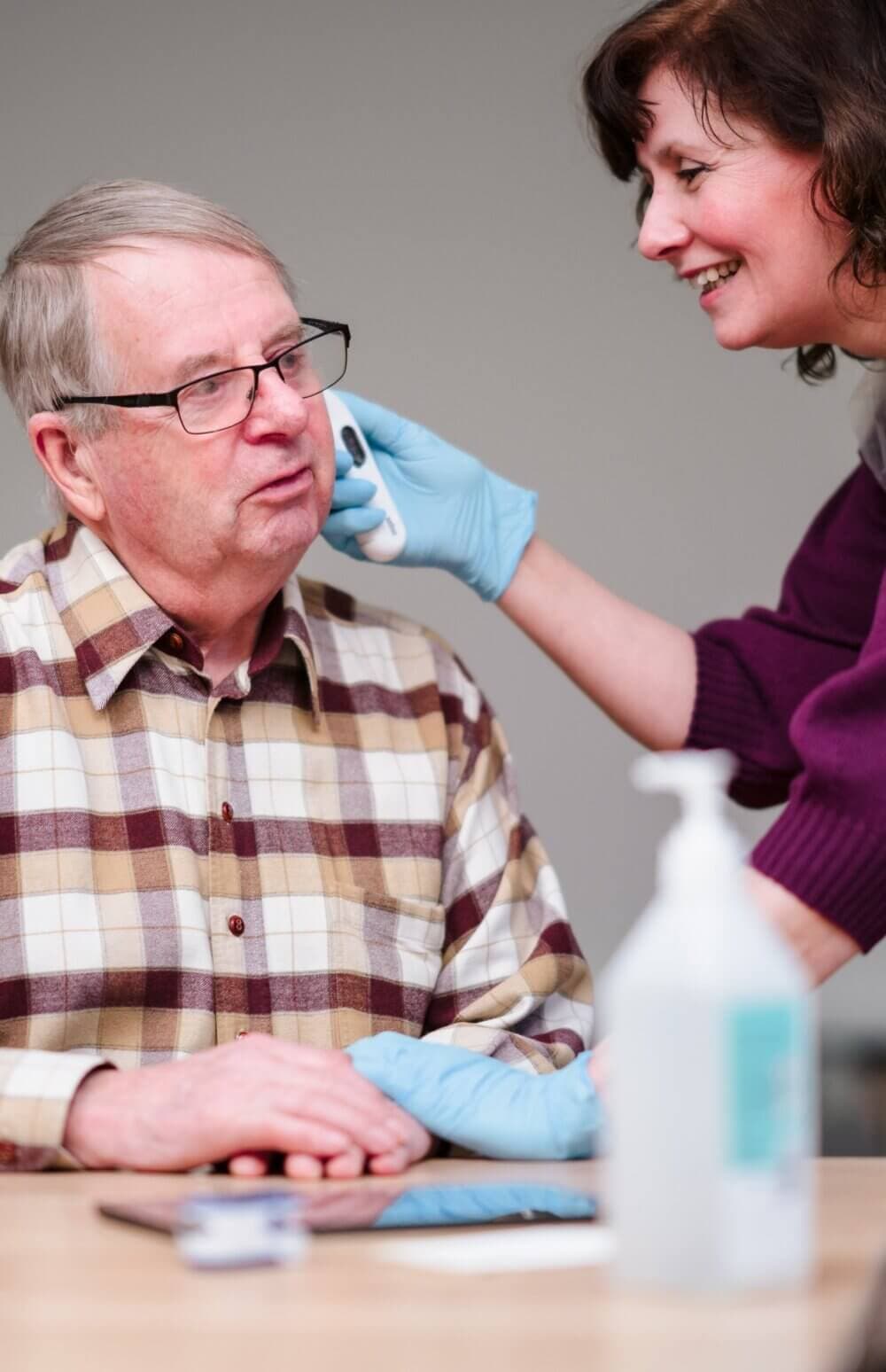 A Care Professional uses an ear thermometer to check the temperature of an elderly man wearing glasses and a plaid shirt. - Home Instead Southampton
