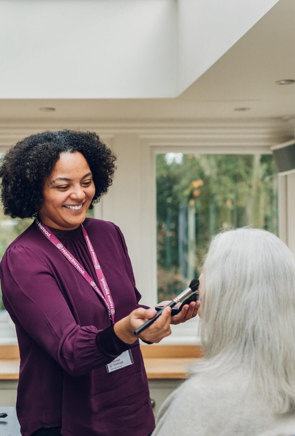 A woman smiling while applying makeup to another person's face in a bright, airy room. - Home Instead