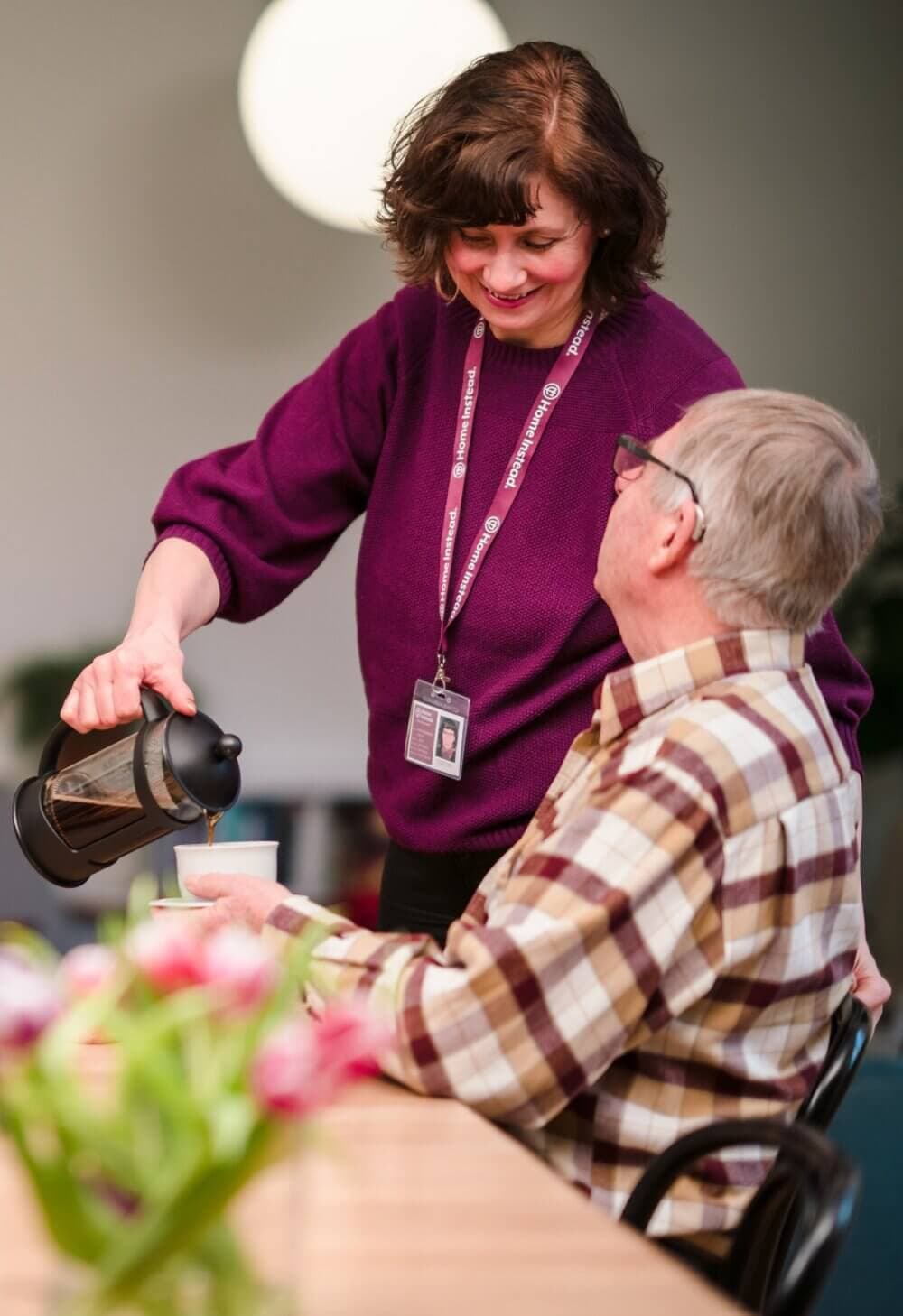 A woman in a purple sweater pours coffee for an older man seated at a table with flowers. - Home Instead