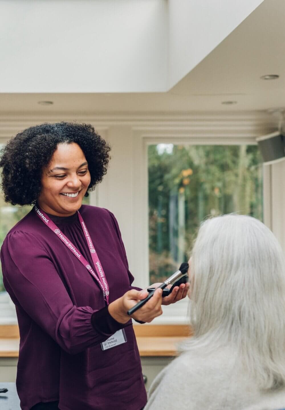 A woman with a name tag applies makeup to an elderly woman in a well-lit room. - Home Instead