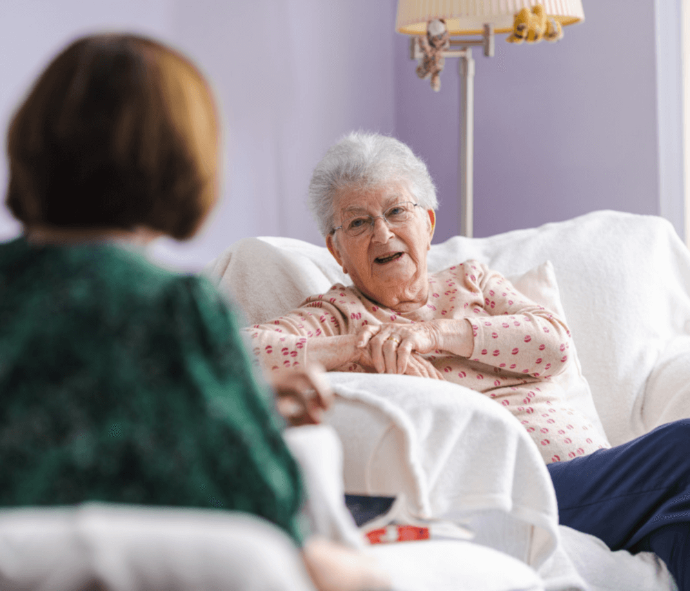 Elderly woman in a pink polka dot shirt sits in a white chair, smiling and conversing with another person in green. - Home Instead