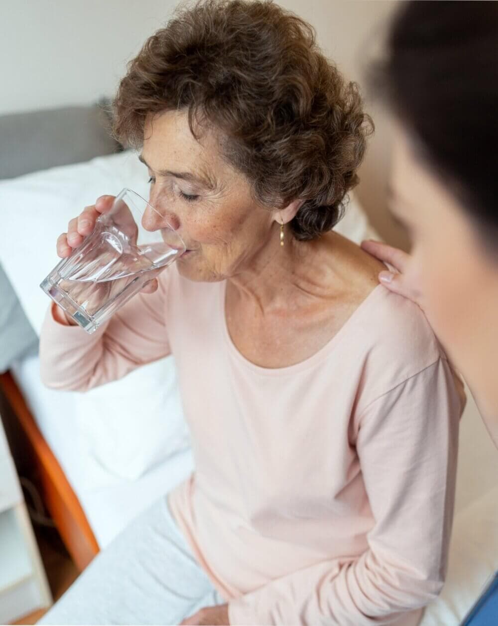 Elderly woman drinking a glass of water while sitting on a bed, with a person gently touching her shoulder. - Home Instead