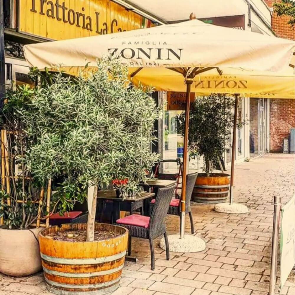 Outdoor cafe with wooden planters, tables, chairs, large umbrellas, and "Trattoria La" sign in background. - Home Instead