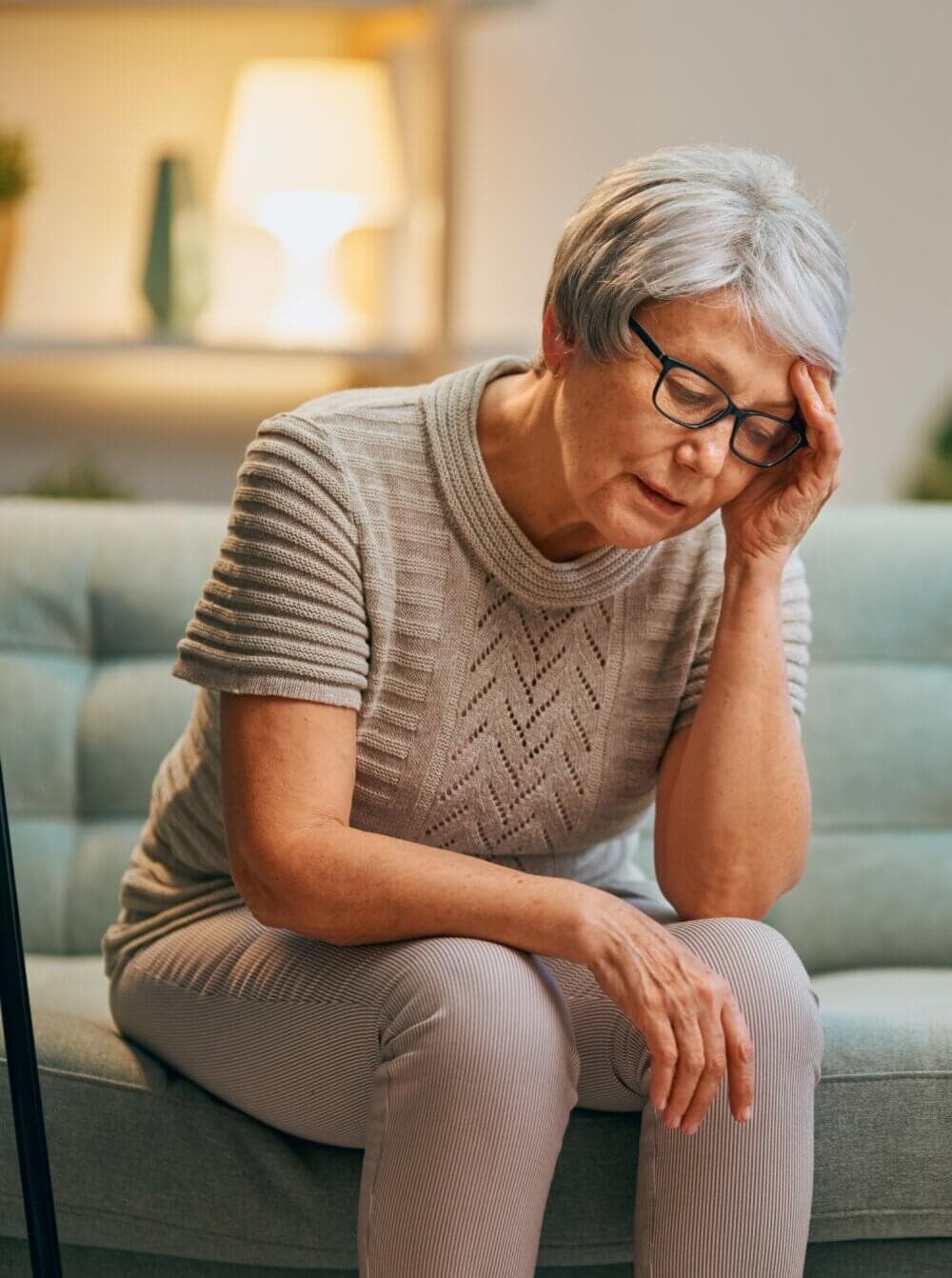 Older woman sitting on a couch, holding her head in one hand, appearing tired or stressed. - Home Instead