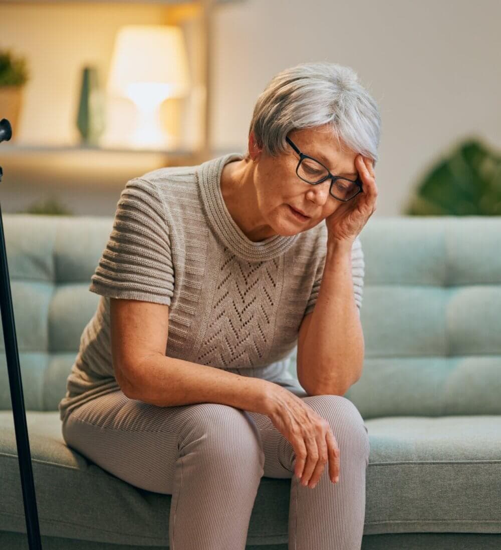 Elderly woman seated on a couch, holding her head with a hand, looking distressed. - Home Instead