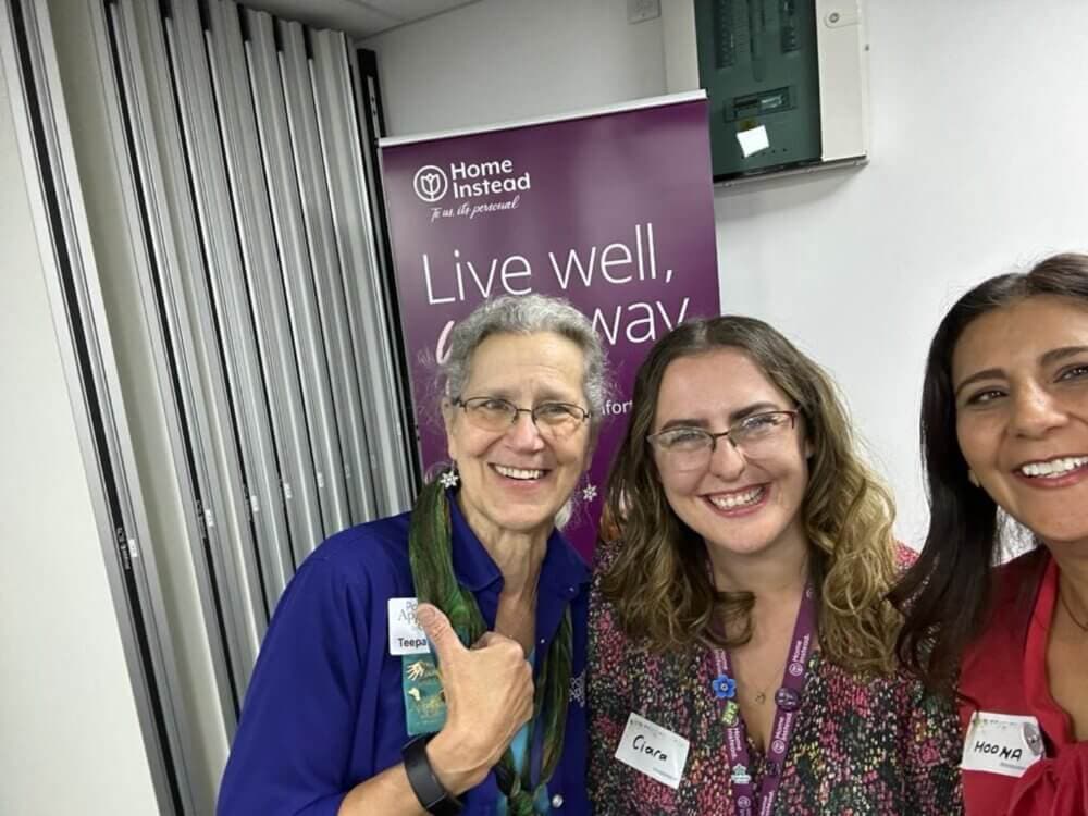 Three women are smiling in front of a banner that reads "Home Instead. Live well, your way. - Home Instead