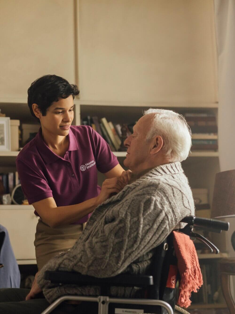 A caregiver in a purple shirt assists an elderly person in a wheelchair with adjusting their gray sweater. - Home Instead