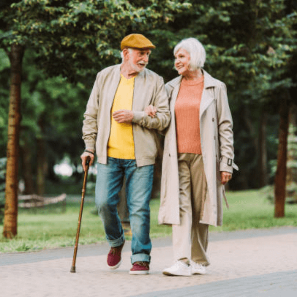 An elderly couple walks arm in arm on a park path, smiling and enjoying the outdoors. The man uses a cane. - Home Instead