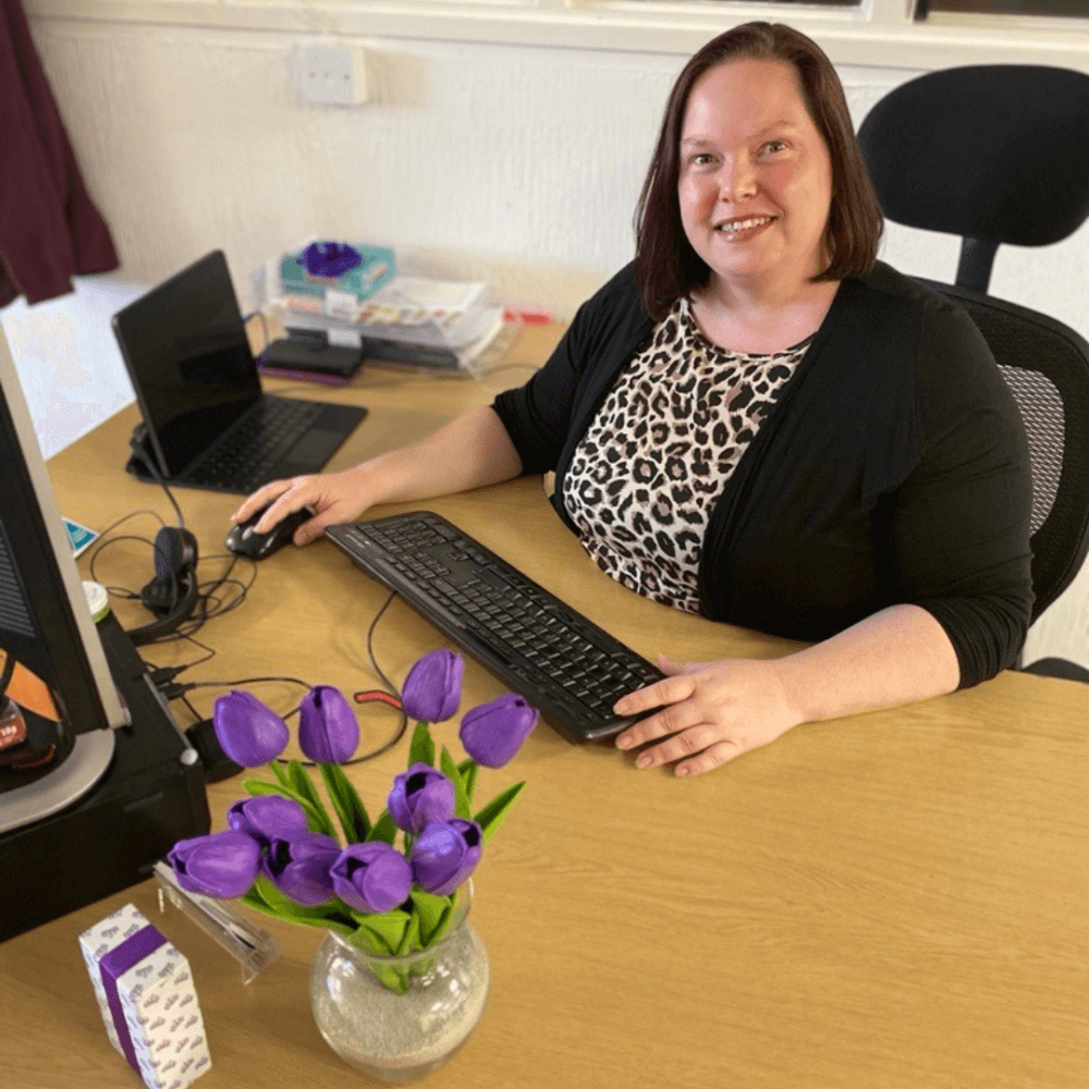 Smiling woman at desk with laptop and keyboard, vase of purple tulips in foreground. Office supplies in background. - Home Instead