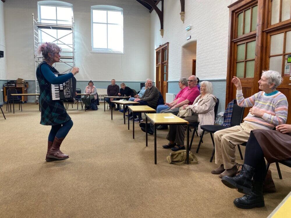 A woman with an accordion conducts a group of seated seniors during a music class in a hall with scaffolding by the windows. - Home Instead