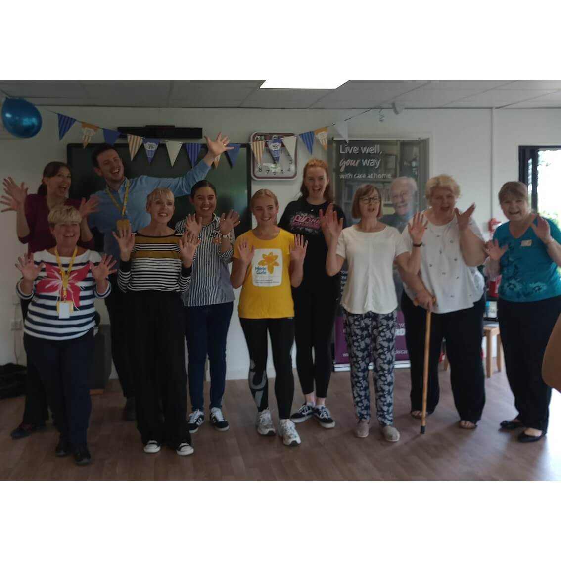 A group of people of various ages are smiling and holding their hands up in a well-lit room decorated with bunting. - Home Instead