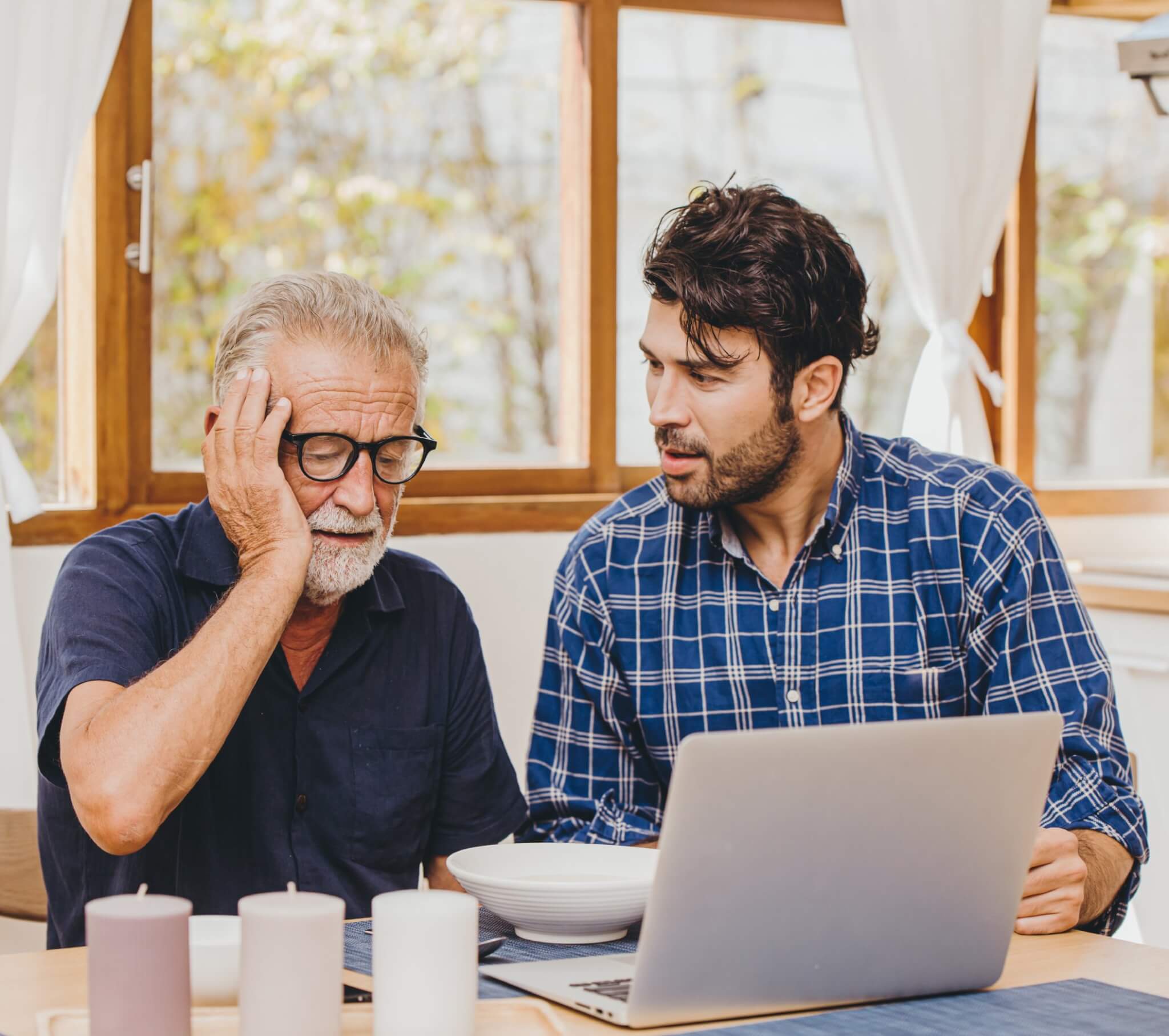 Older man and younger man looking concerned while sitting at a table with a laptop and candles. - Home Instead