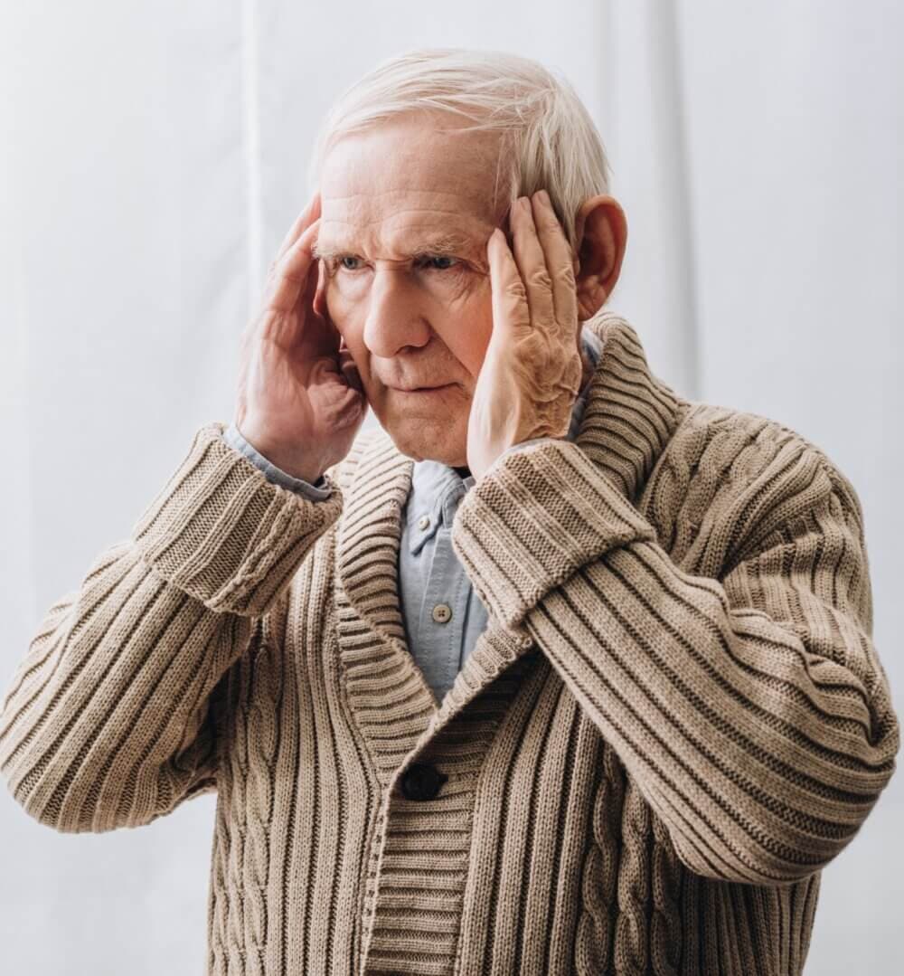 Elderly man wearing a beige knitted cardigan holding his head, looking concerned. - Home Instead