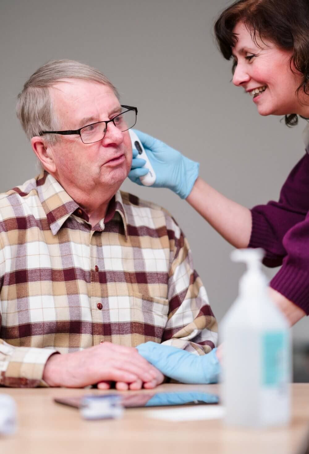 A caregiver wearing blue gloves wipes the face of an elderly man with glasses in a checkered shirt. - Home Instead