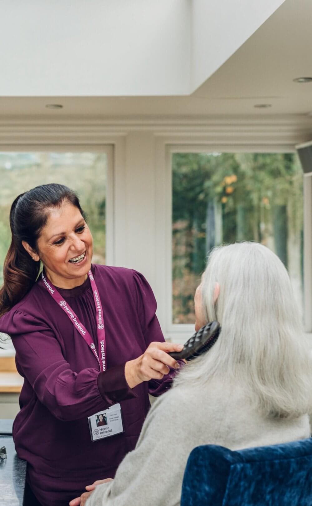 A woman in a purple shirt, wearing a name badge, is brushing the hair of a seated elderly woman with gray hair. - Home Instead