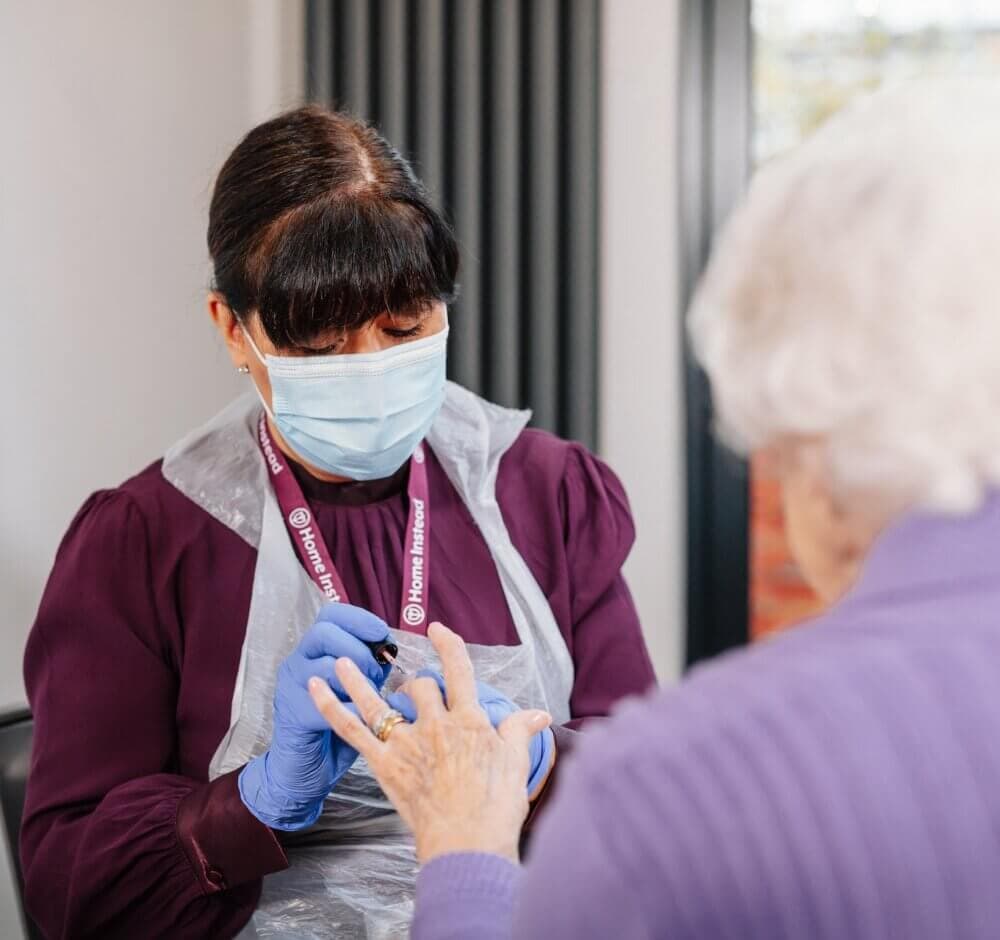 A caregiver with a name badge smiling as they assist an elderly person with brushing their hair in a sunny room. - Home Instead