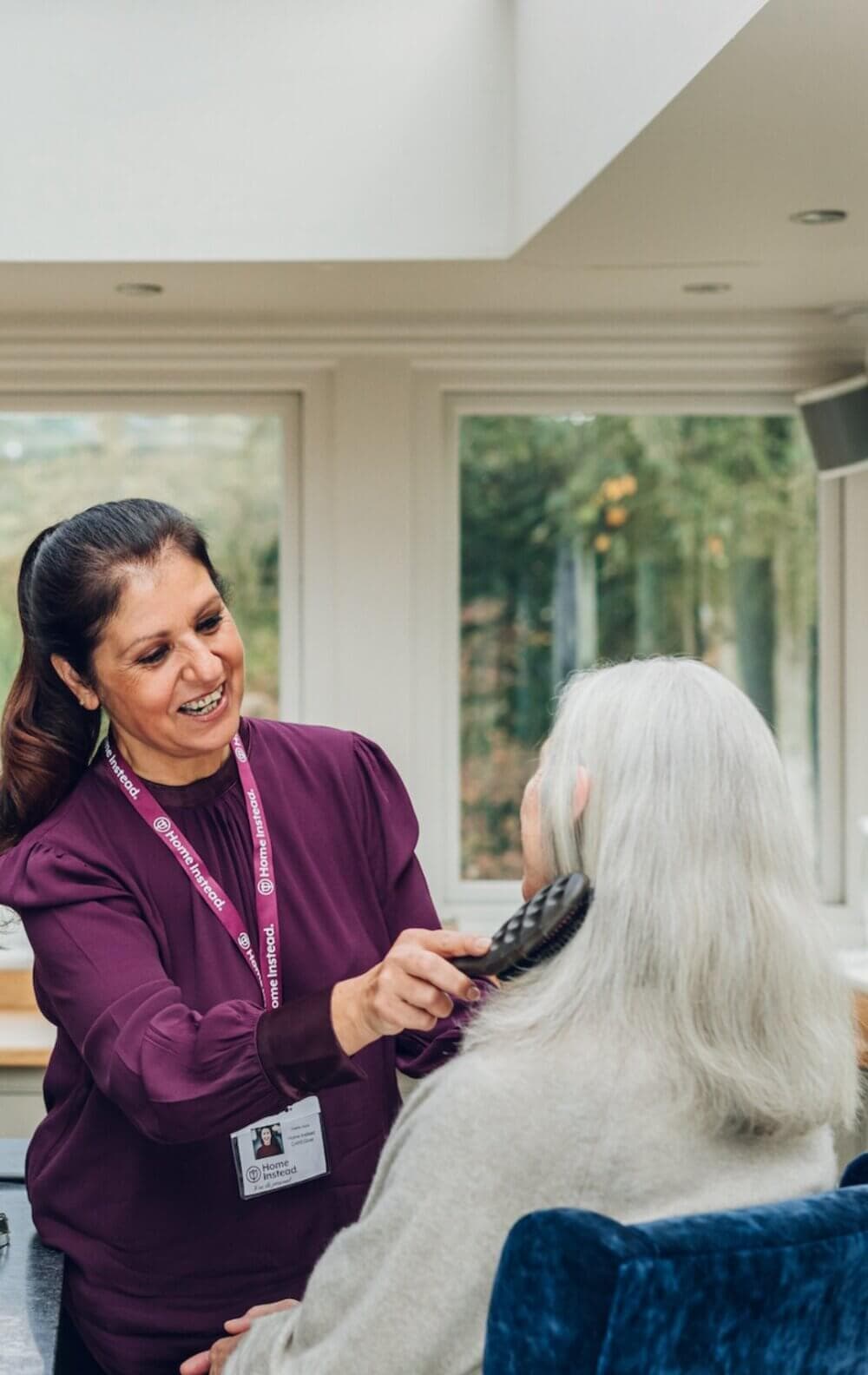 A woman in a purple top brushes the white hair of an elderly person sitting in a chair in a bright room. - Home Instead
