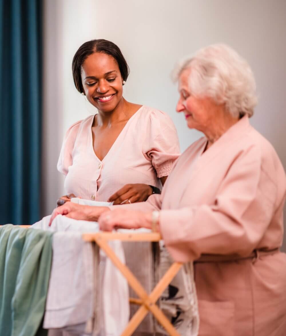 A caregiver smiles while brushing an older woman's hair in a well-lit room with large windows. - Home Instead