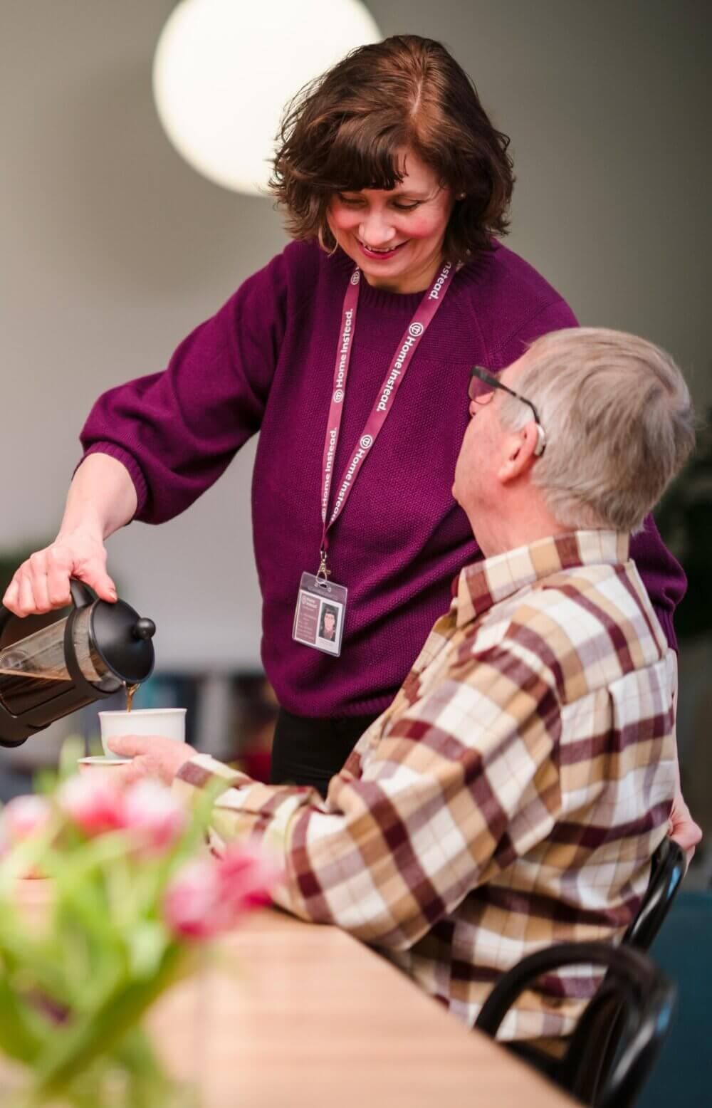 A smiling woman in a purple sweater pours coffee for an older man seated at a table. - Home Instead