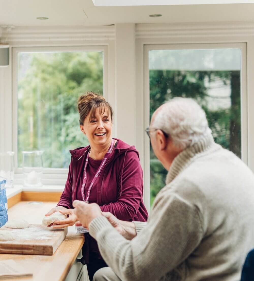 A woman and an elderly man smile while sitting at a table in a bright room with large windows. - Home Instead