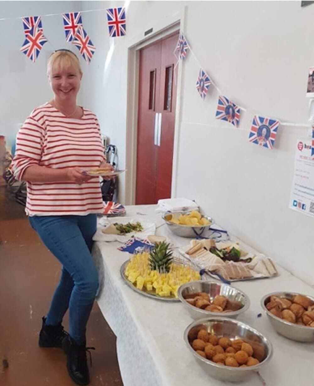 Woman smiling next to a table with various food items, decorated with British flag bunting. - Home Instead
