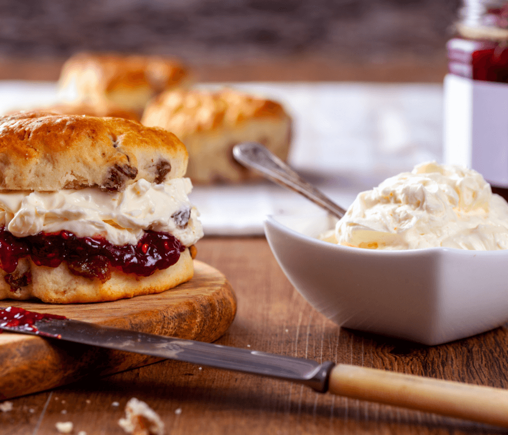 A scone with cream and jam on a wooden board, with a dish of cream and a knife in the foreground. - Home Instead