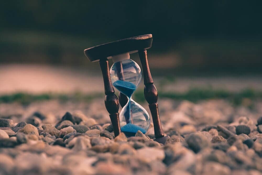 An hourglass with blue sand tilted on a bed of small pebbles with a blurred natural background. - Home Instead