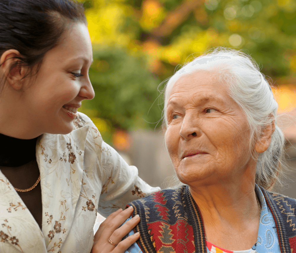 A smiling woman and elderly woman looking at each other outdoors, with the younger one's hand on the elder's shoulder. - Home Instead