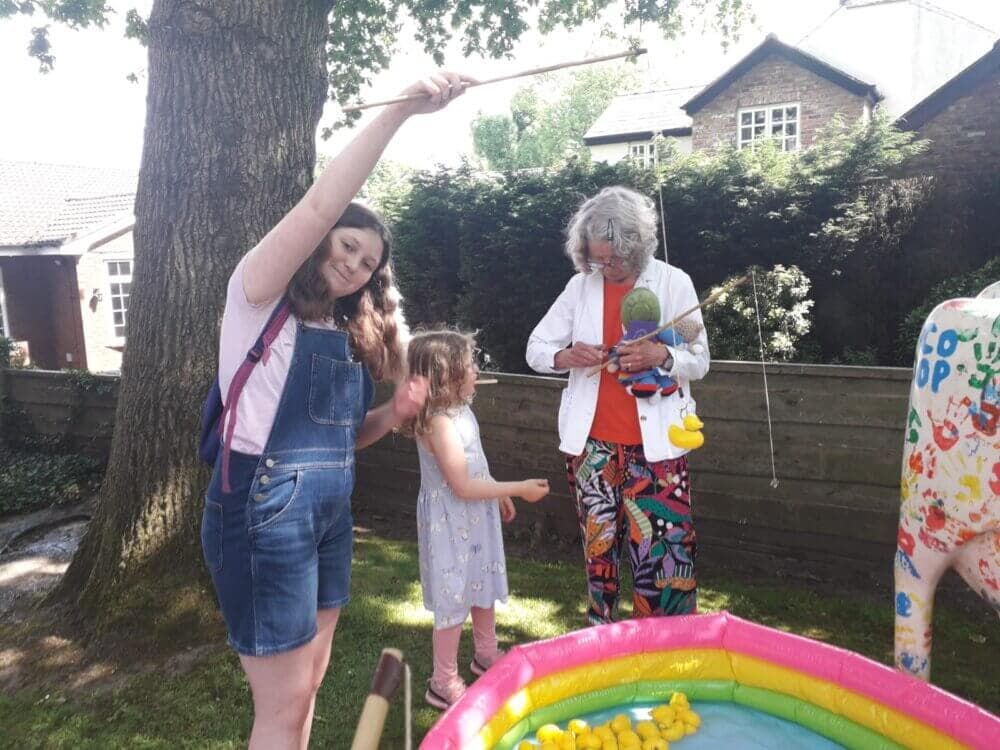 Three people playing a fishing game with rubber ducks next to an inflatable pool in a garden on a sunny day. - Home Instead