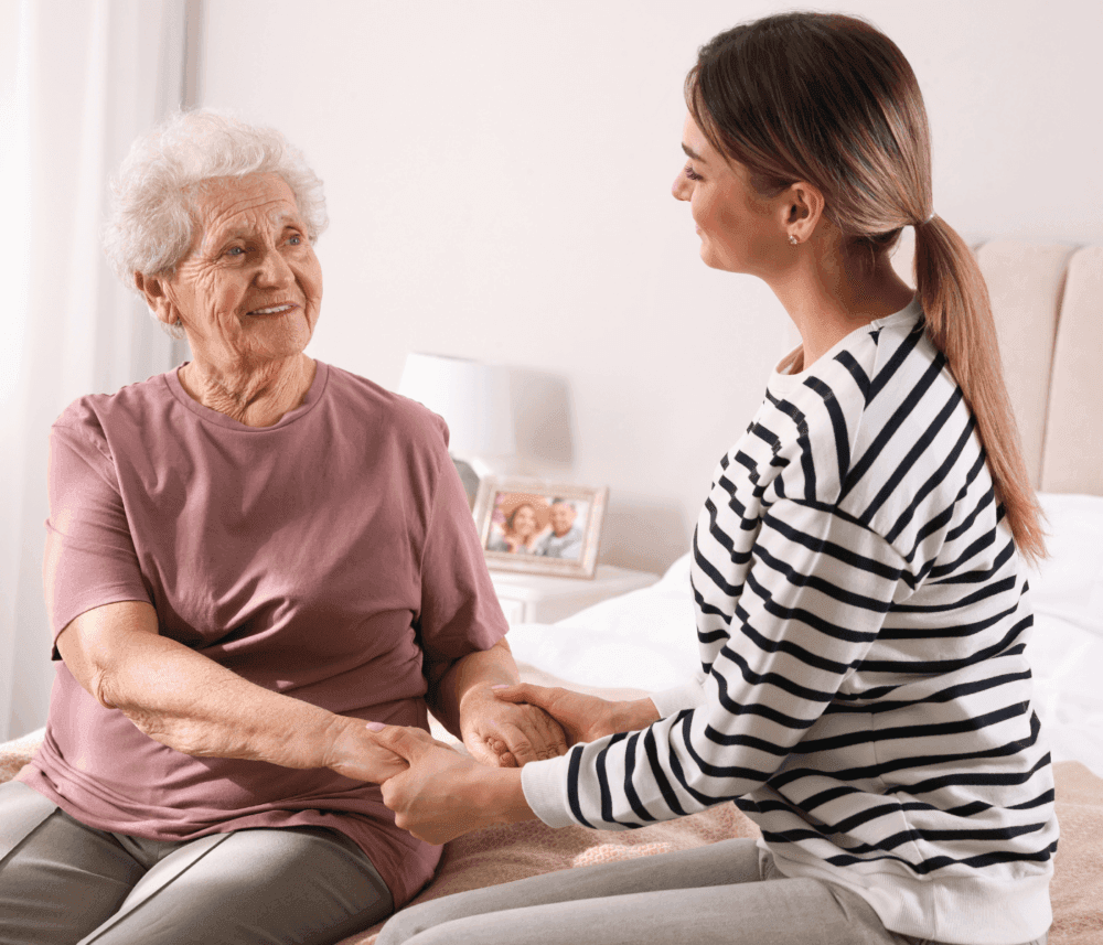 A young woman holds hands with an elderly woman, both sitting on a bed in a cozy room, smiling warmly at each other. - Home Instead