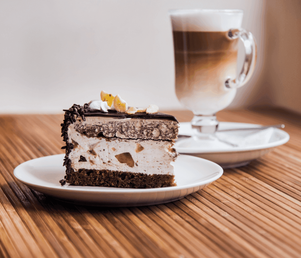 Slice of layered chocolate and cream cake on a plate next to a glass of layered coffee with foam on a wooden table. - Home Instead
