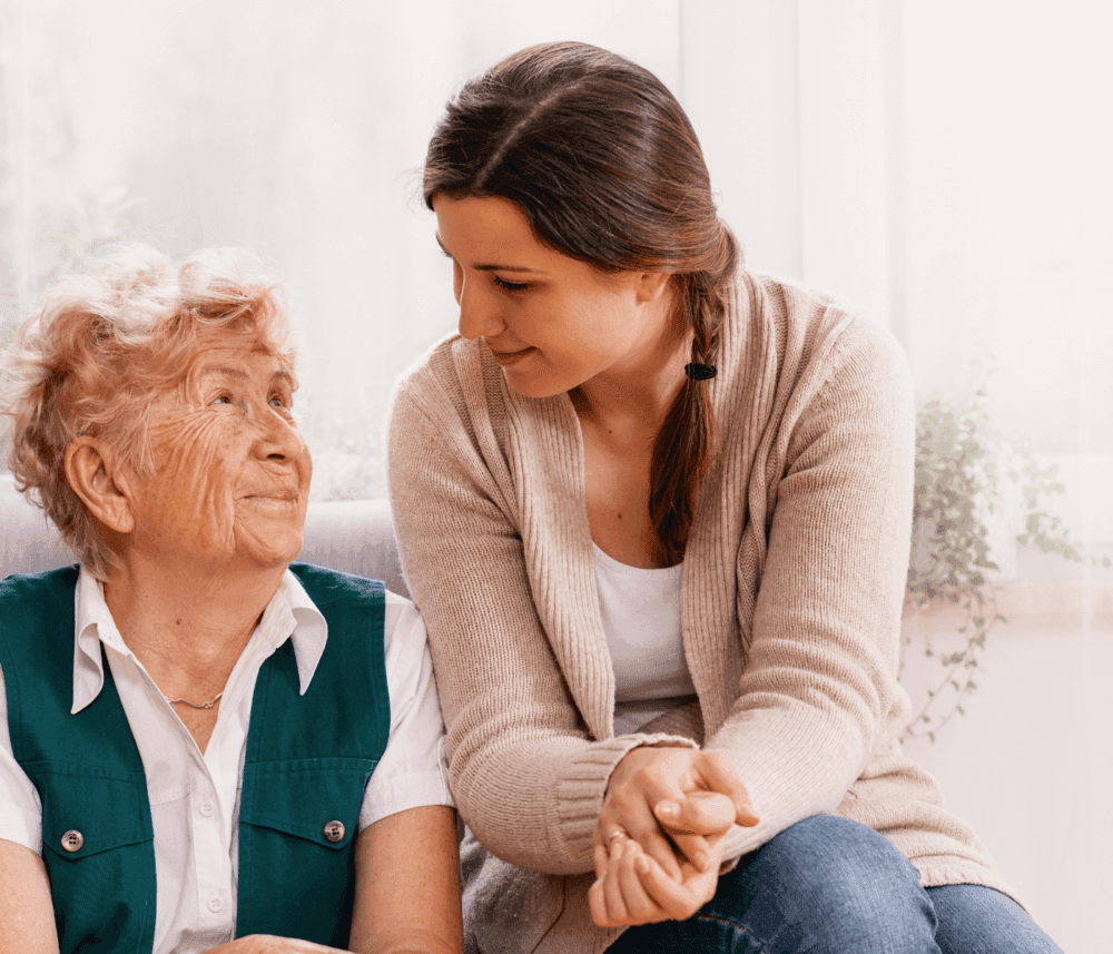 An elderly woman with a younger woman sitting together, sharing a tender moment and smiling warmly at each other. - Home Instead