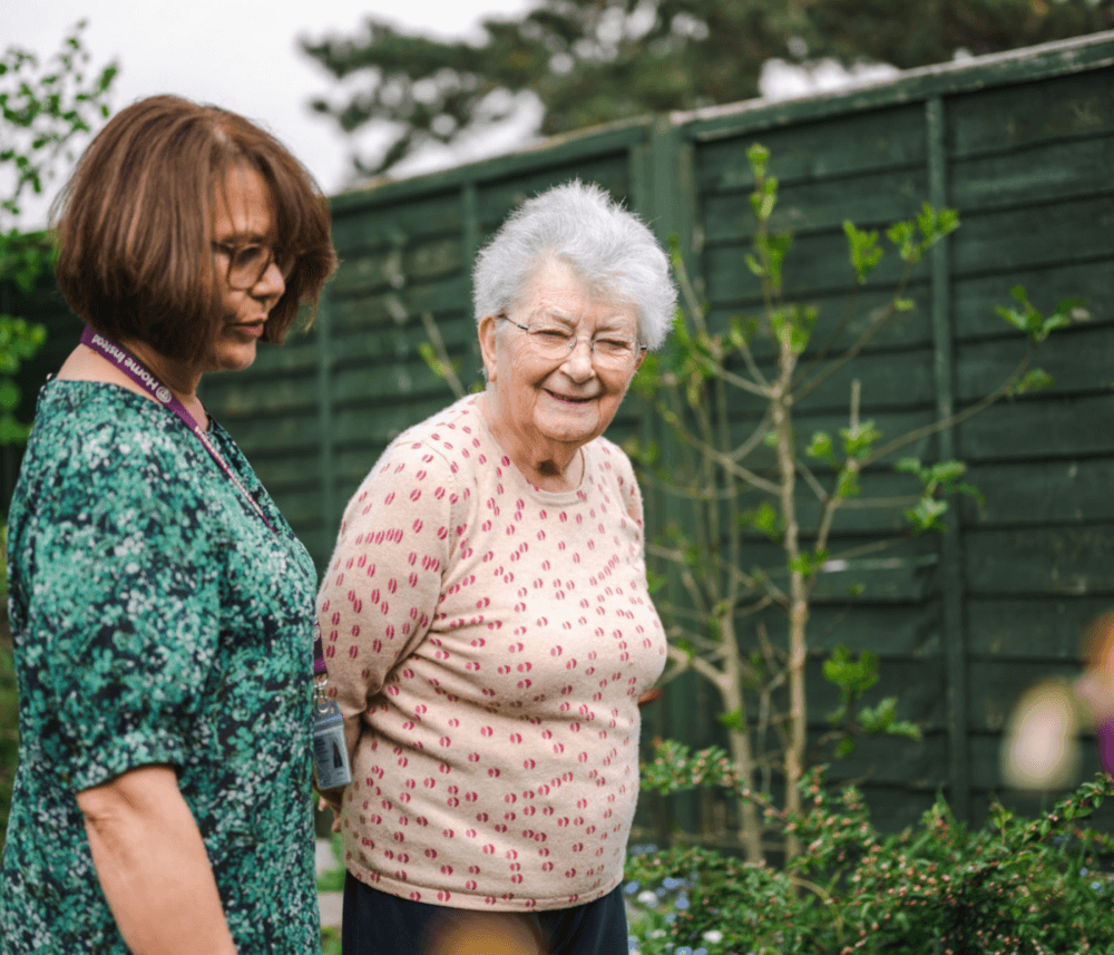 Two women, an elderly woman in a garden, and a younger woman beside her, walking together near a green fence. - Home Instead