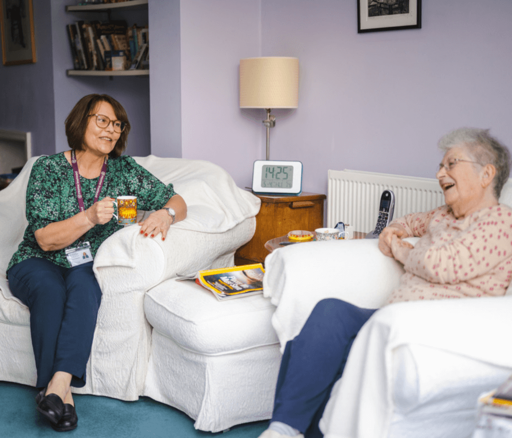Two women sit and chat in a cozy living room. One holds a mug, and the clock on the table shows 14:25. - Home Instead