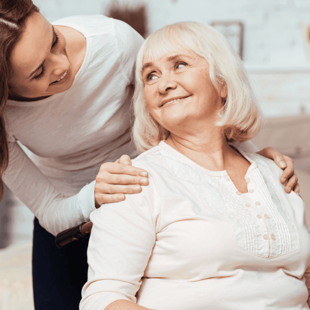 A young woman smiles and places her hand on an older woman's shoulder, who is seated and smiling back at her. - Home Instead