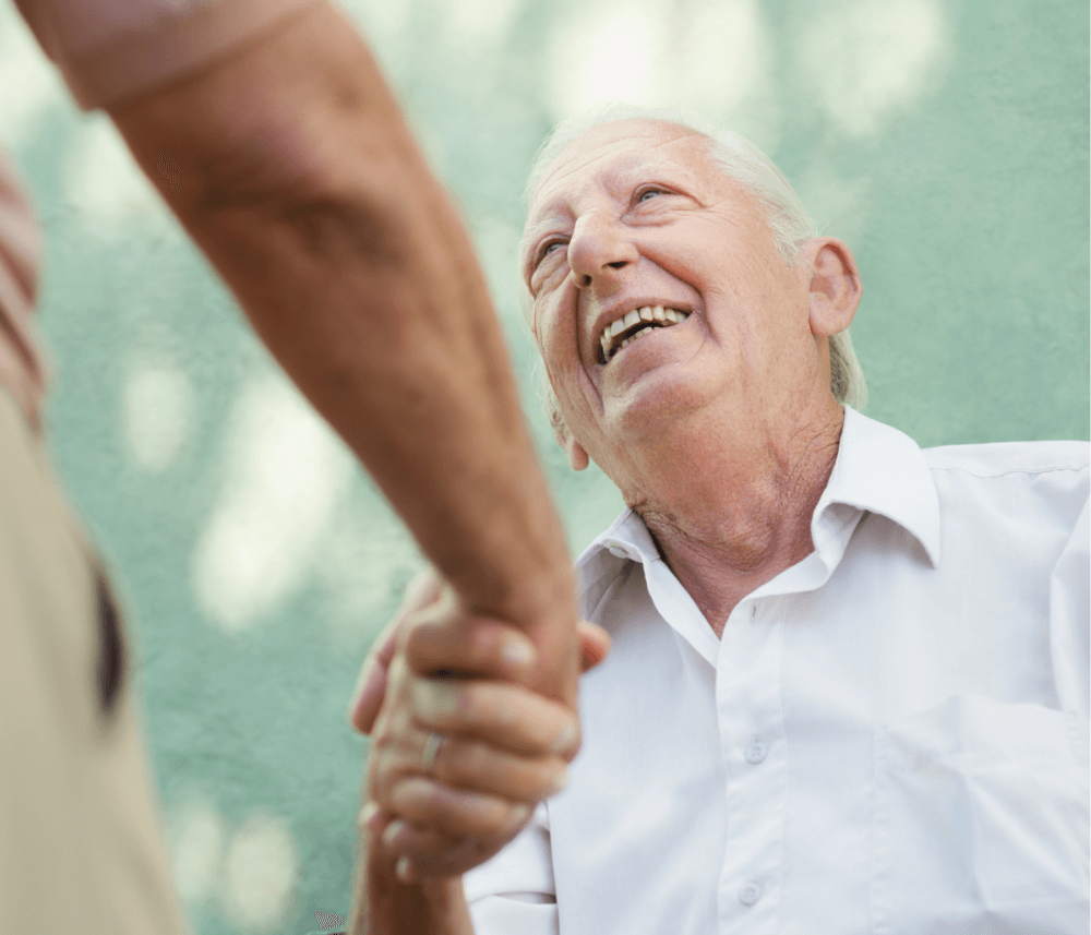 Elderly man in a white shirt smiling and shaking hands with another person. Background is a blurred green outdoor setting. - Home Instead