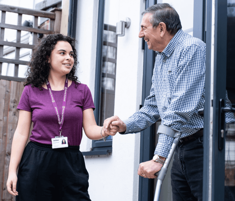 A young woman assists an elderly man with a crutch at a doorway, both smiling warmly at each other. - Home Instead