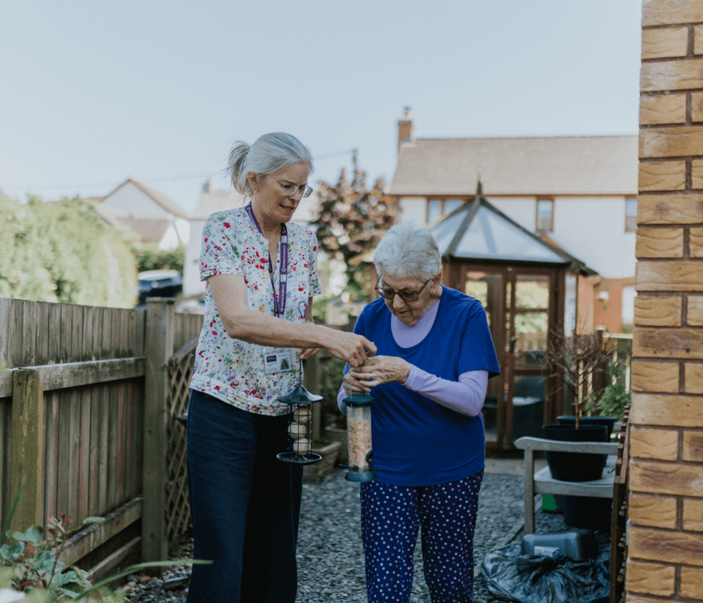 Two elderly women enjoying an outdoor activity involving bird feeders beside a house with a garden shed in the background. - Home Instead