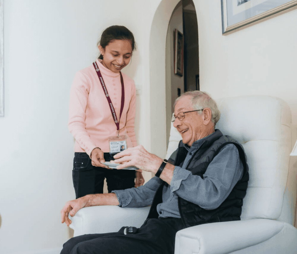 Caregiver handing a cup to a seated elderly man, both smiling inside a bright, warmly lit room. - Home Instead