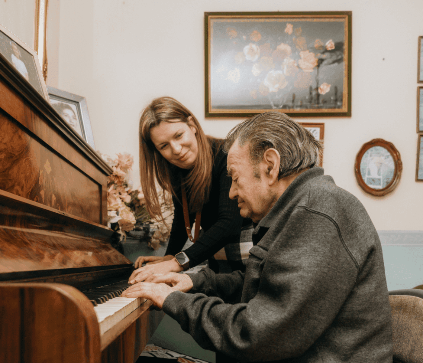 An elderly man plays the piano while a woman smiles and assists him in a room with framed pictures on the wall. - Home Instead