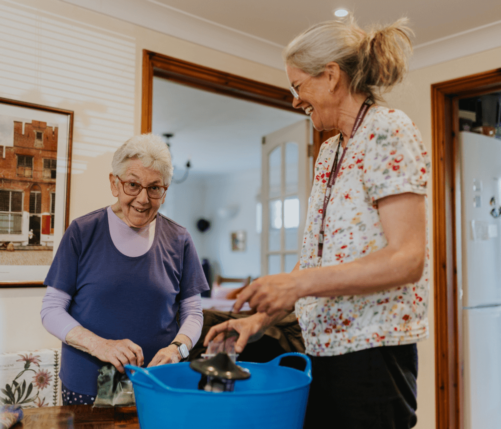 Two women smile while doing housework together in a cozy kitchen, with a blue basin on the table in front of them. - Home Instead