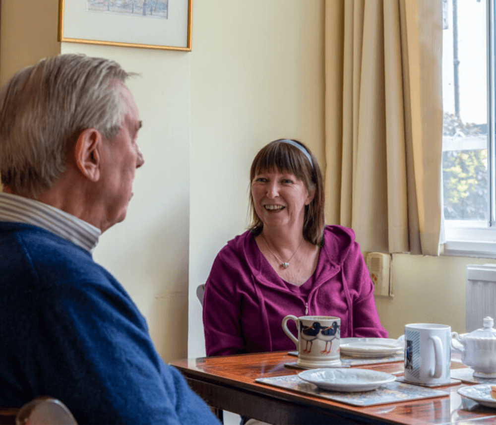 An older man and a woman smile and talk at a dining table with cups and plates in a sunlit room. - Home Instead