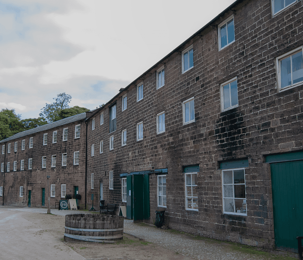 A long, brick building with many windows and green doors, set against a partly cloudy sky with a wooded area nearby. - Home Instead