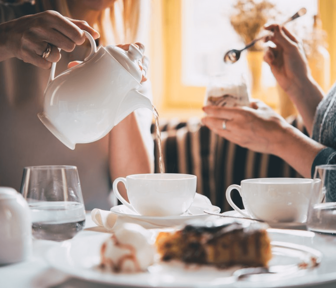 Two people enjoying tea, one pouring from a white teapot, with pastries and glassware on a table in a cozy setting. - Home Instead