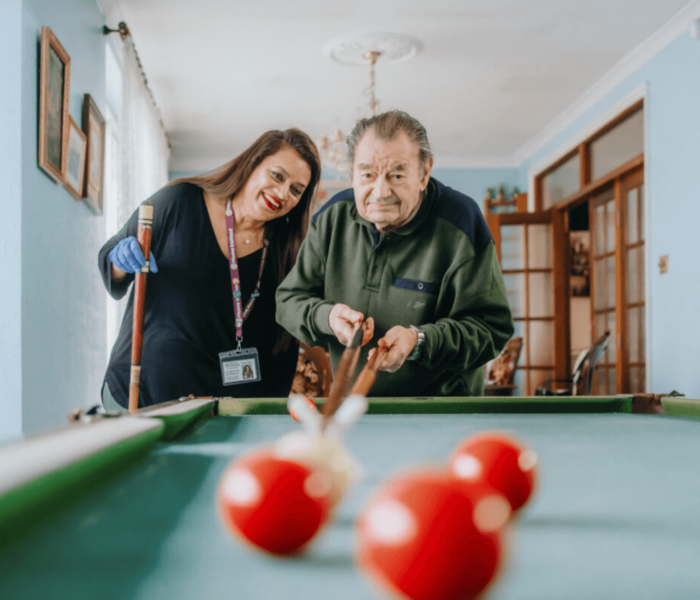 A man aims to hit pool balls while a woman assists him in a well-lit room. - Home Instead