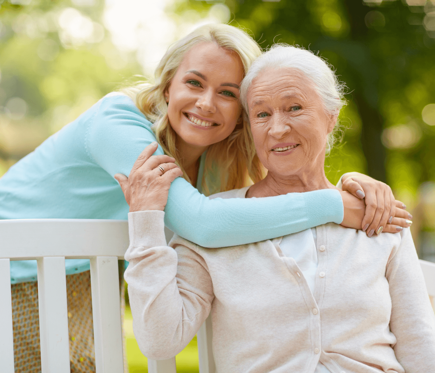 A young woman hugs an elderly woman from behind as they sit on a bench outdoors, both smiling warmly. - Home Instead