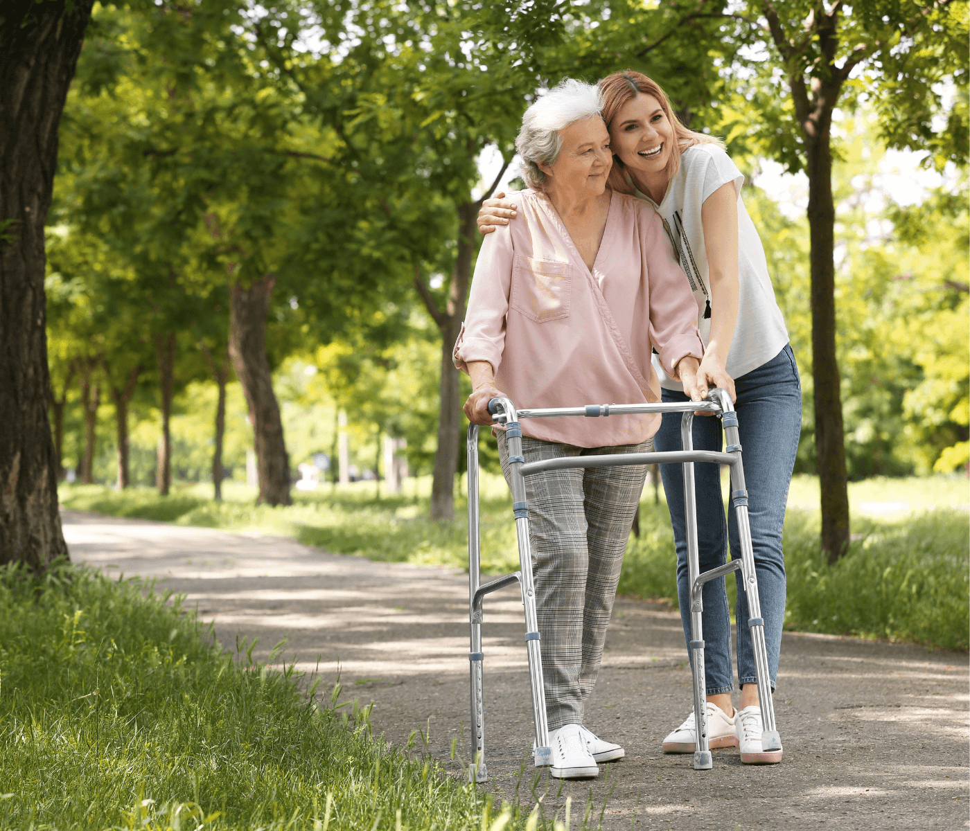 A young woman smiles and hugs an elderly woman using a walker, as they walk together in a park. - Home Instead