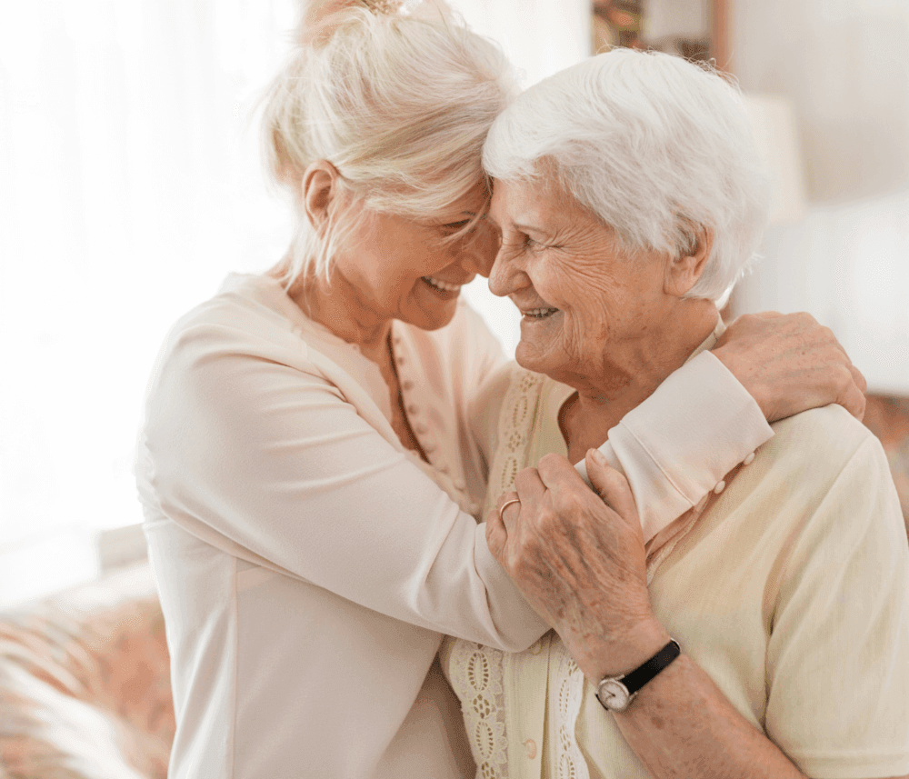 Two elderly women smiling and sharing an affectionate hug in a warmly lit room. - Home Instead