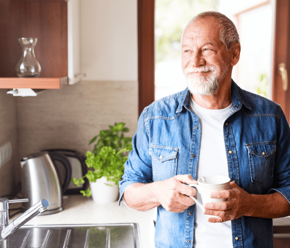 Smiling elderly man with a beard holding a mug in a kitchen, standing near a sink with a kettle and plants in the background. - Home Instead